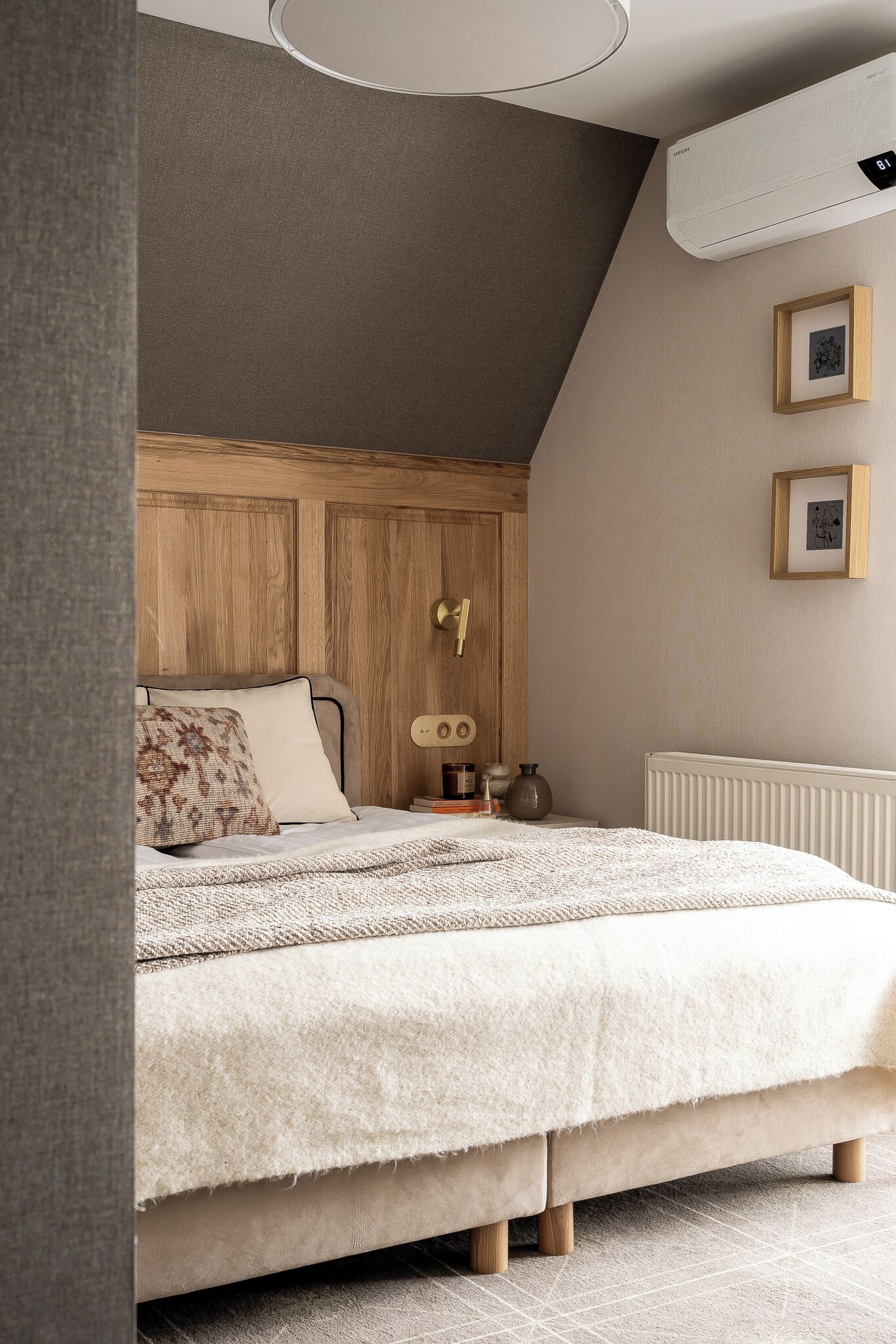 Un dormitorio con cabecera de madera, cama con edredón blanco, mesita de noche con lámpara y radiador.