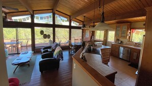 A spacious living area with a wooden ceiling, a dining area with a table and chairs, a kitchen with a stove and sink, and large windows offering views of greenery.