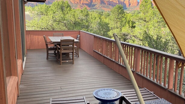 A wooden deck with a table and chairs, a blue table, and a view of red rock formations and green trees.