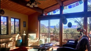 A cozy room with a wooden ceiling, a large window with a view of mountains, a comfortable chair, a small table, and a sofa.