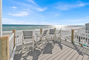 Eine Holzterrasse mit weißen Stühlen und einem Geländer mit Blick auf einen Strand.