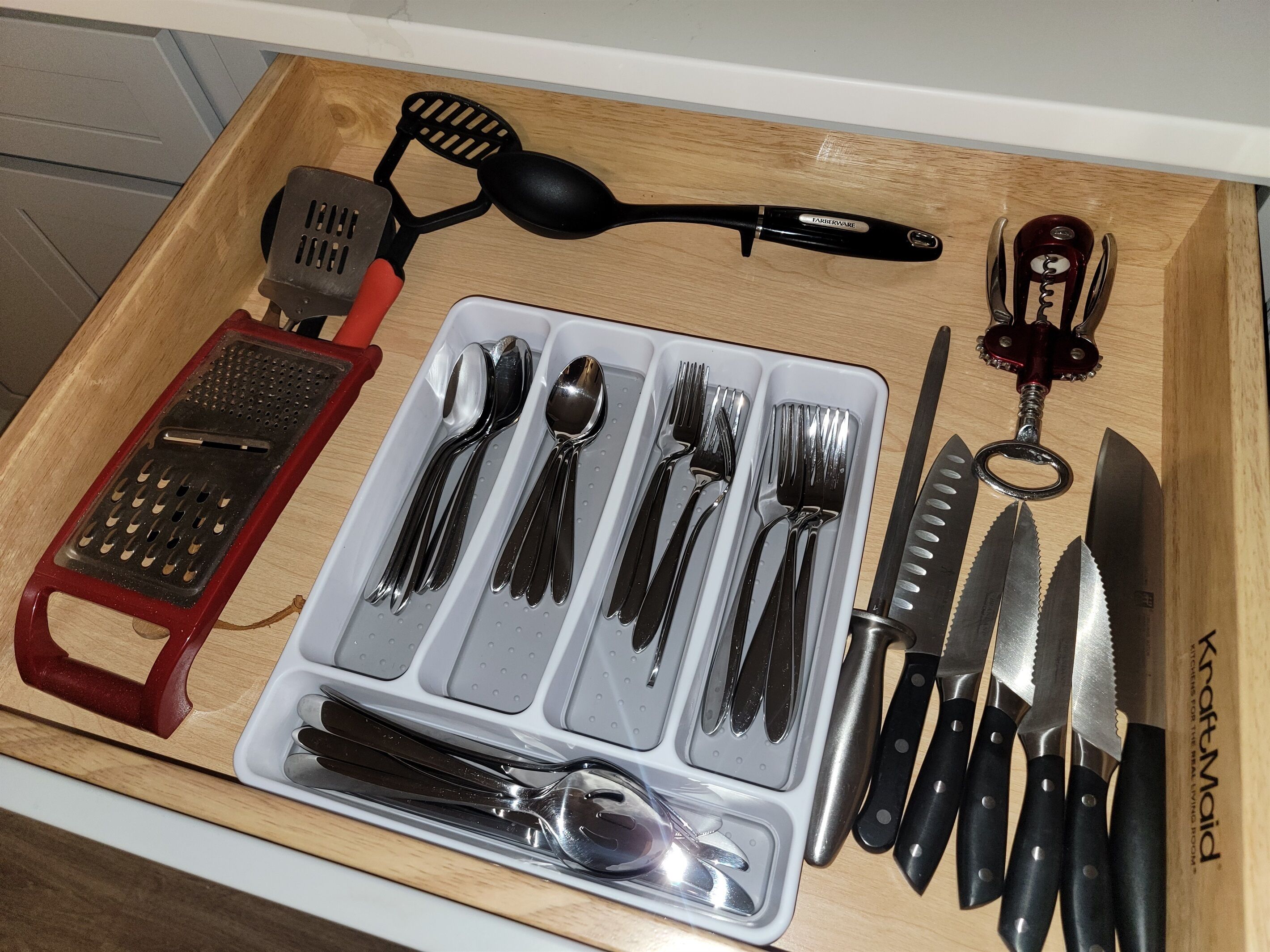 A kitchen drawer with utensils including a grater, spatula, spoon set, forks, knives, and a corkscrew.