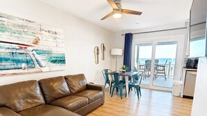 A living room with a brown leather sofa, a ceiling fan, a dining area with teal chairs, and a view of the ocean through large glass doors. - 11A - 1BR $700+ in freebies incl golf, On Beach (Miramar Beach)