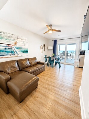 A living room with a brown leather sofa, a ceiling fan, a dining area with teal chairs, and a view of the ocean through large glass doors.