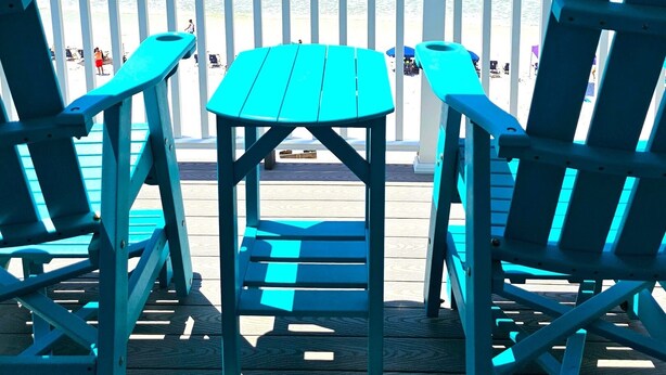 Una terrazza con due poltrone Adirondack color turchese e un tavolino, con vista su una spiaggia, le onde e un cielo azzurro limpido.