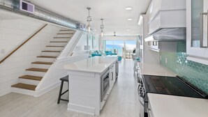 A modern kitchen with a central island, a staircase with wooden steps, and a view of the ocean. - Miramar Beach 2BR • Beach + Free Beach Setup & Fun (Miramar Beach)