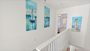 A hallway with white wainscoting and a white railing. There are three paintings on the walls: two of them depict sailboats on a blue water background, and one shows a colorful beach scene with a lifebuoy. - Miramar Beach 2BR • Beach + Free Beach Setup & Fun (Miramar Beach)