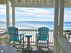 Ein überdachter Außenbereich mit zwei Stühlen und einem Tisch, mit Blick auf einen Strand und das Meer im Hintergrund.