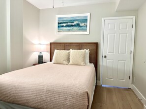 Ein Schlafzimmer mit einem Bett, einem Nachttisch, einer Lampe, einer Tür und einem gerahmten Bild an der Wand.