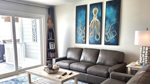 A living room with a brown leather sofa, a wooden coffee table, and a blue patterned rug. There are three abstract paintings on the wall, a bookshelf, and a lamp on a side table. - 7B - 2BR $700+ in freebies incl golf, On Beach (Miramar Beach)