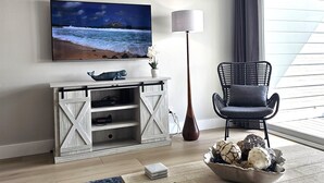 A living room with a TV mounted on the wall, a wooden entertainment unit, a floor lamp, a chair, and a window with curtains. - 7B - 2BR $700+ in freebies incl golf, On Beach (Miramar Beach)