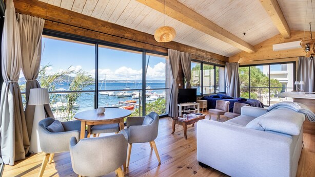 A spacious living room with a large window overlooking a marina, wooden ceiling beams, and a cozy seating area with a sofa and chairs.