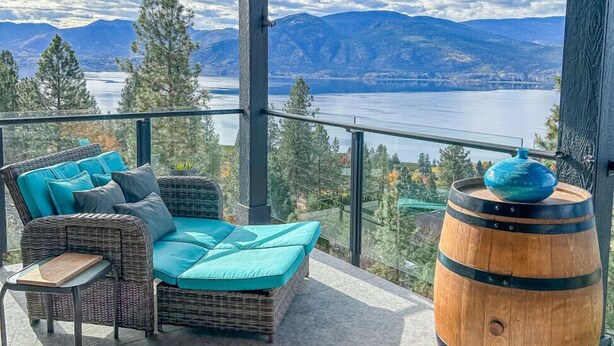 Ein Balkon mit einer Rattan-Couch, einem Holzfass und Blick auf einen See und Berge.