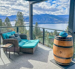 A balcony with a wicker sofa, a wooden barrel, and a view of a lake and mountains. - Naramata Hillside Luxe Retreat (Naramata)