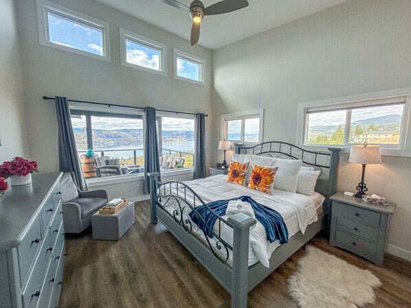 A bedroom with a bed, a dresser, a chair, a nightstand, a lamp, and a view of the outdoors. - Naramata Hillside Luxe Retreat (Naramata)