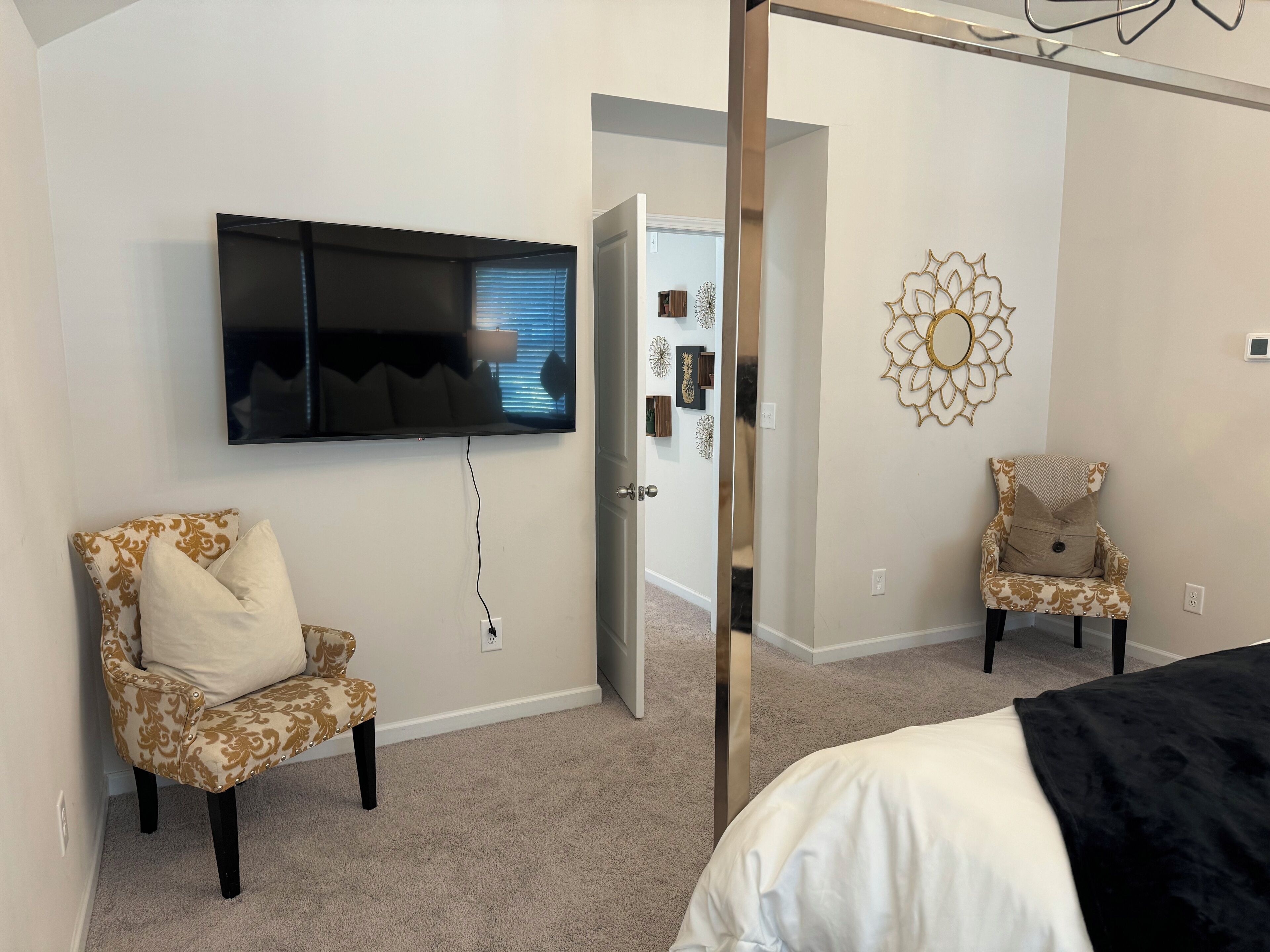 A bedroom with a TV mounted on the wall, two patterned armchairs, a decorative wall piece, and a bed with a black cover.