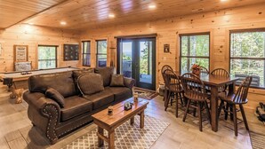 Ein gemütliches Blockhaus-Interieur mit Holzbalkendecke, Billardtisch, Ledersofa, Esstisch mit Stühlen und großen Fenstern mit Blick ins Freie.