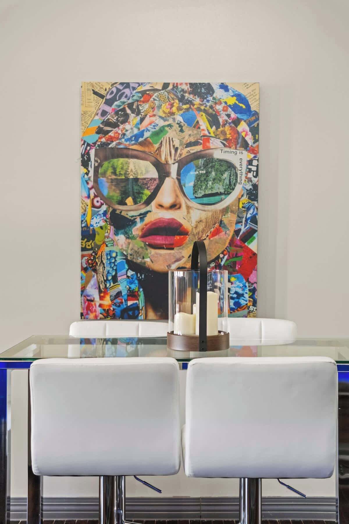 A dining area with a glass table and white chairs, a colorful abstract painting on the wall, and a candle holder on the table.