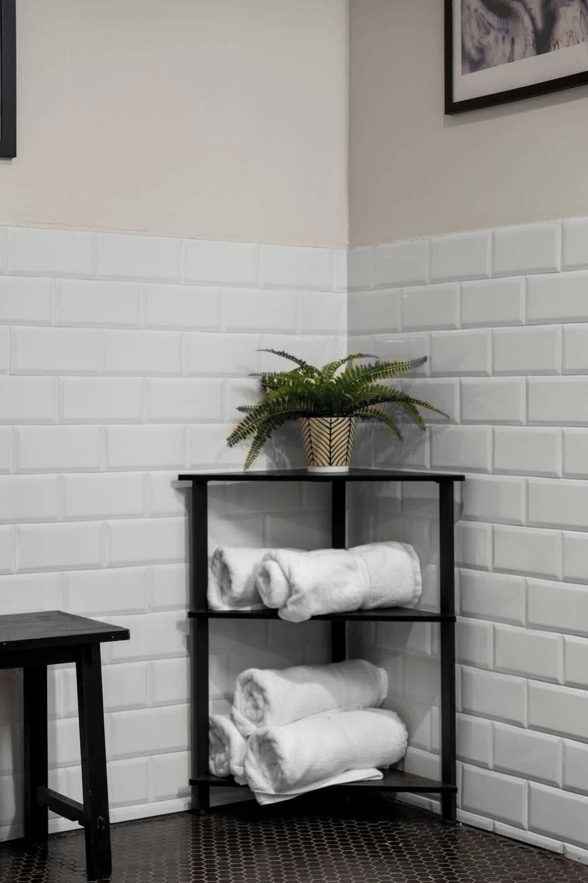 A corner with a black metal shelf holding rolled towels and a potted plant.