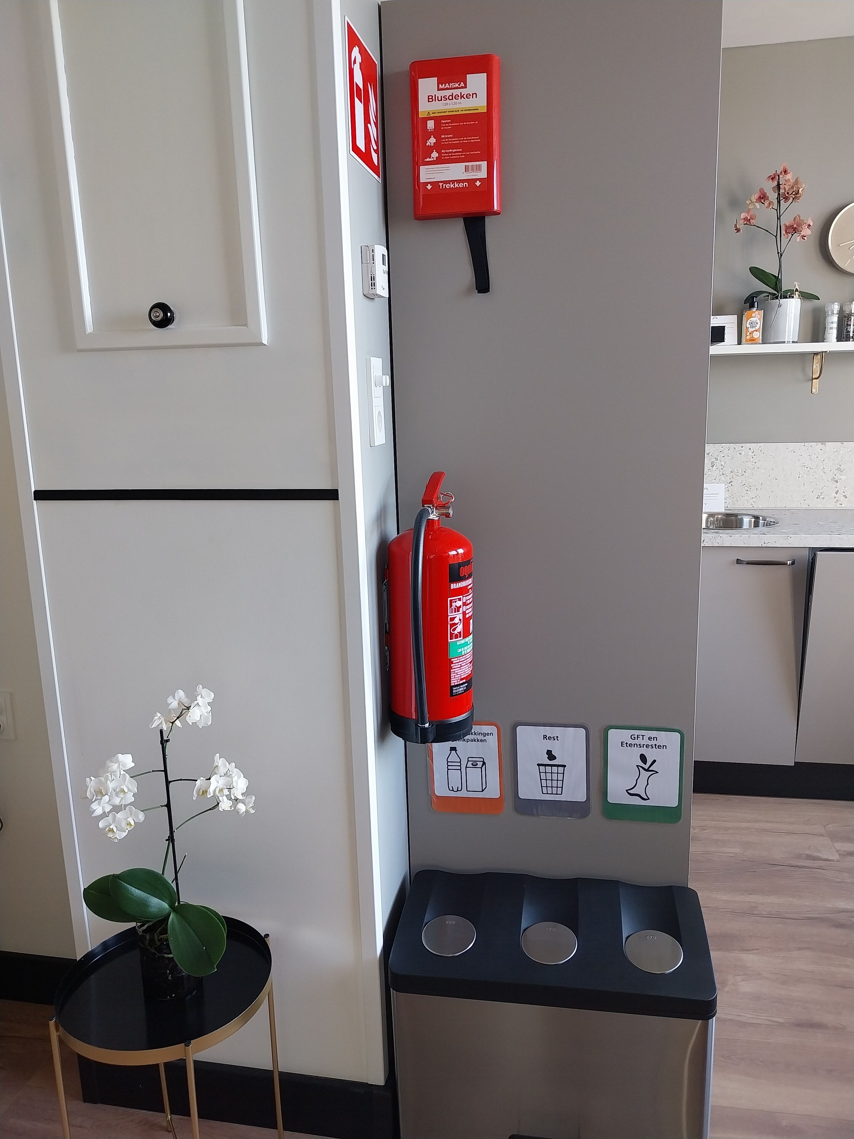 Een kamer met een deur, een aan de muur bevestigde brandblusser, een prullenbak en recyclingbakken.