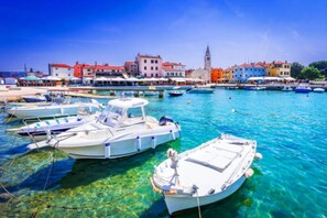 Une ville côtière aux eaux turquoise et limpides, où des bateaux sont amarrés le long de la plage, et où des bâtiments aux couleurs vives se dressent, avec une tour imposante en arrière-plan.