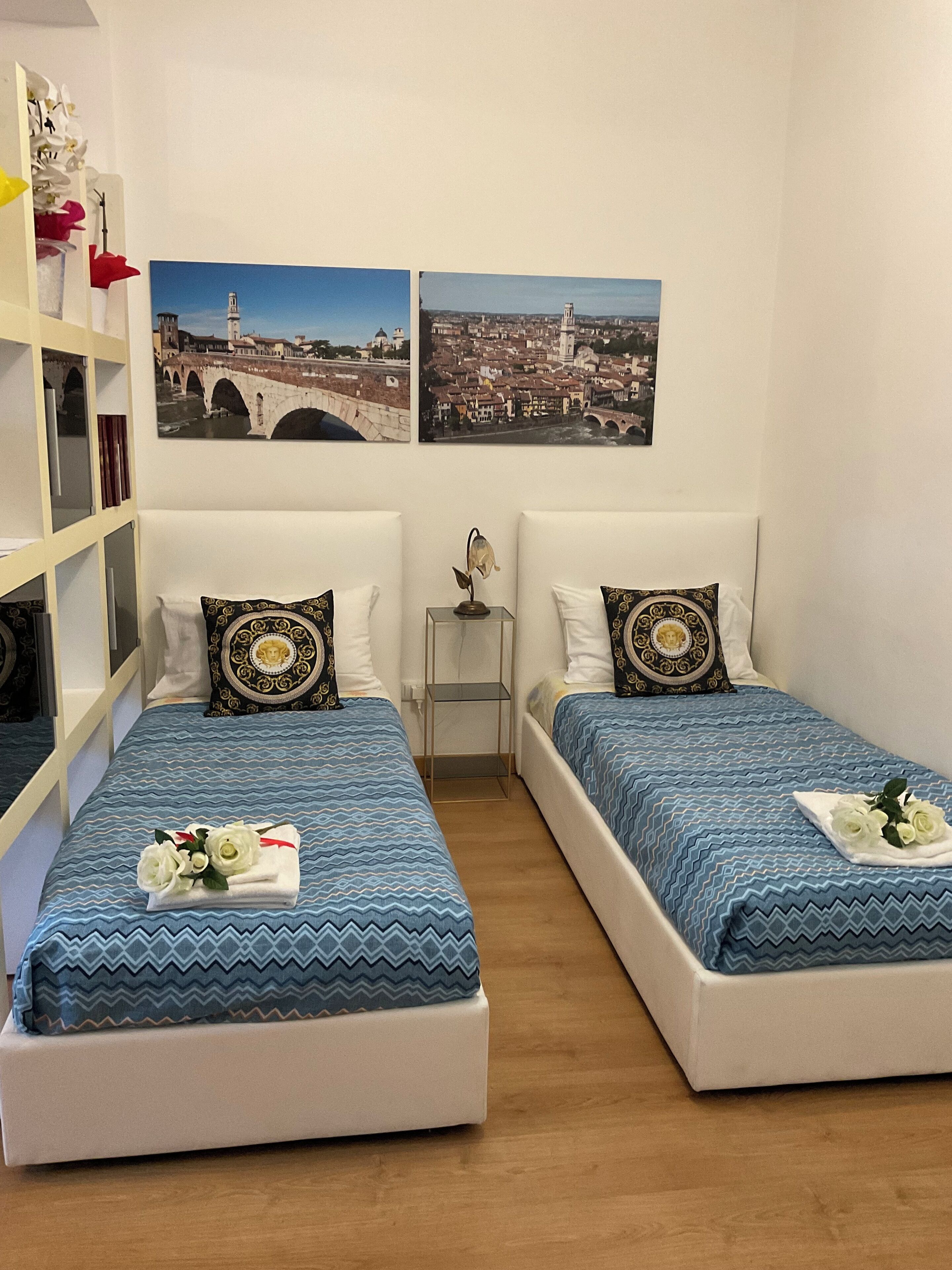 Une chambre d’hôtel avec deux lits, chacun recouvert d’une couvre-lit à motifs bleus et agrémenté de coussins blancs. Deux tableaux encadrés ornent le mur, ainsi qu’une petite table de chevet avec une lampe et une bibliothèque contenant divers objets.