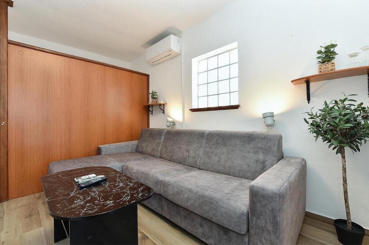 A modern living room with a grey sofa, a small round coffee table, a wooden wardrobe, a window with blinds, and a potted plant.