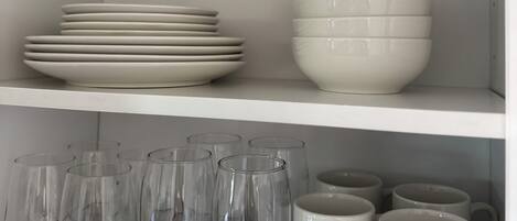 A white shelving unit with stacked plates, bowls, and clear glassware.