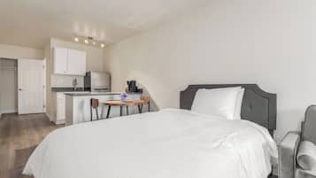 A hotel room with a bed, a small desk with a chair, a kitchenette, and a white door.