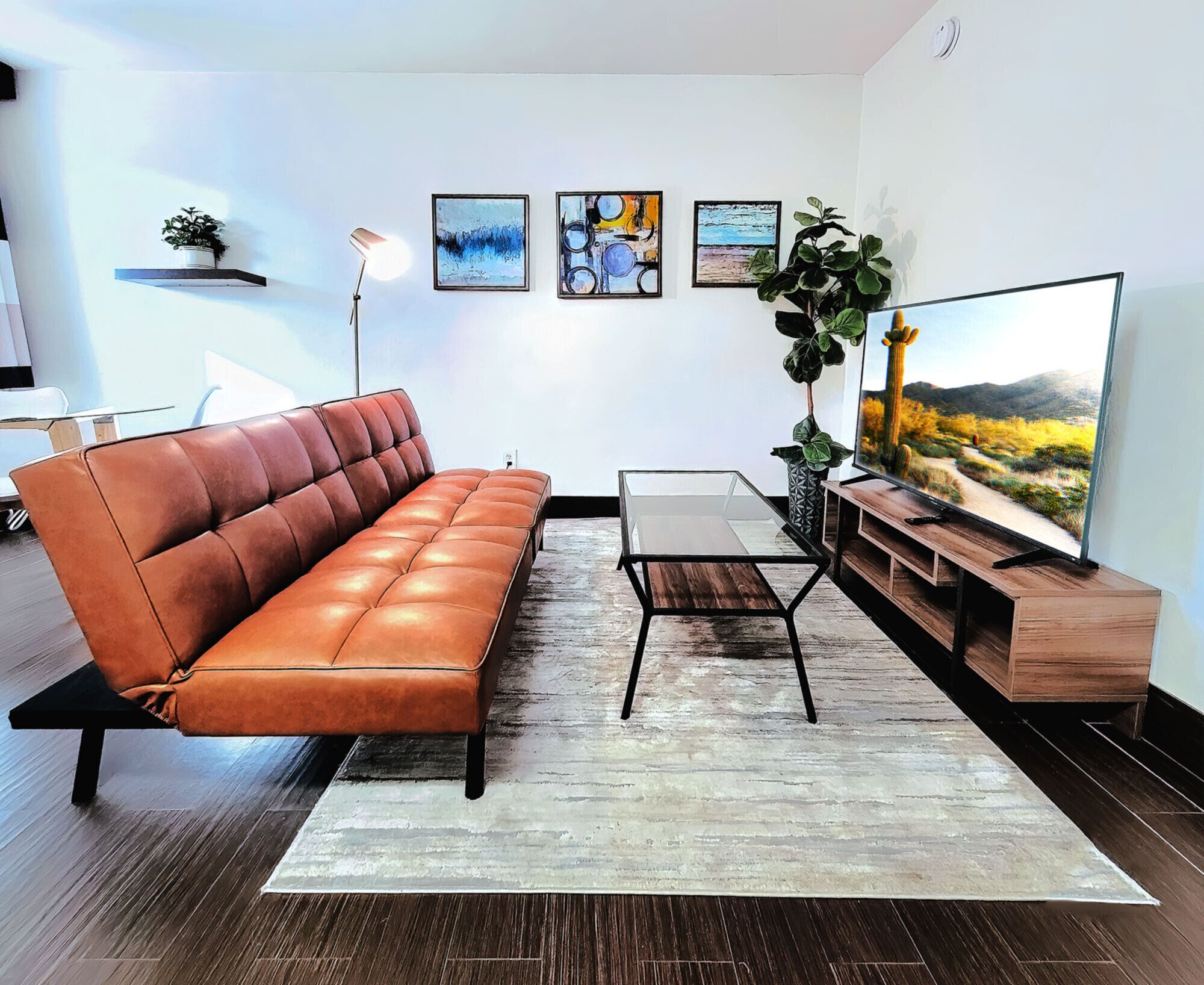 A modern living room with a leather sofa, a glass coffee table, a wooden TV stand, and a large flat-screen TV displaying a desert landscape.