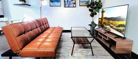 A modern living room with a leather sofa, a glass coffee table, a wooden TV stand, and a large flat-screen TV displaying a desert landscape.