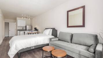 Un dormitorio moderno con una cama, un sofá gris, dos mesas redondas de madera y una zona de cocina al fondo.
