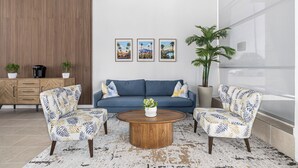 Un salon moderne avec un canapé, deux fauteuils, une table basse et une grande fenêtre. Trois tableaux encadrés ornent le mur, ainsi que deux plantes en pot.