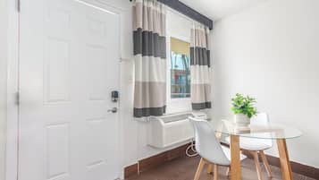Habitació amb una porta blanca, una finestra amb cortines a ratlles, un radiador, una taula de vidre amb cadires i una planta en test.