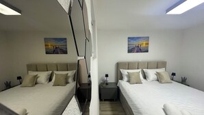 Ein Hotelzimmer mit zwei Betten, jeweils mit Kopfteil, weißer Bettwäsche und einem Nachttisch mit Lampe. An den Wänden hängen zwei Gemälde, und eine Deckenleuchte ist zu sehen.