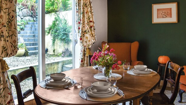 A dining area with a table set for four, a vase with flowers, and a view of a garden through a glass door.