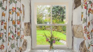 A window with a floral curtain, a vase with flowers, and a view of a garden.