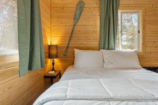 A wooden cabin bedroom with a bed, a nightstand, a lamp, and a window with curtains. - Peaceful Cabin on Whitefish Chain (Crosslake)