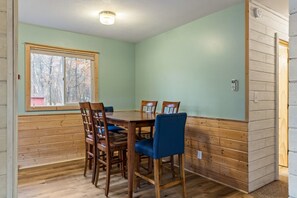 A dining area with a wooden table and chairs, a window with a view of trees, and a light switch on the wall.