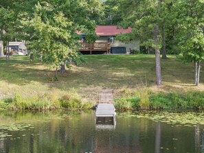 A wooden dock extending into a calm body of water with lily pads, surrounded by trees and a house with a red roof in the background. - Peaceful Cabin on Whitefish Chain (Crosslake)
