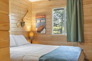 A wooden cabin room with a bed, a poster, a lamp, and a window with curtains. - Peaceful Cabin on Whitefish Chain (Crosslake)