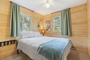 A wooden-paneled bedroom with a bed, two windows, a ceiling fan, and a wall-mounted lamp. - Peaceful Cabin on Whitefish Chain (Crosslake)