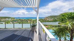 Eine Holzterrasse mit weißem Pergola, mit Blick auf einen See, ein Schwimmbecken und die Küste.