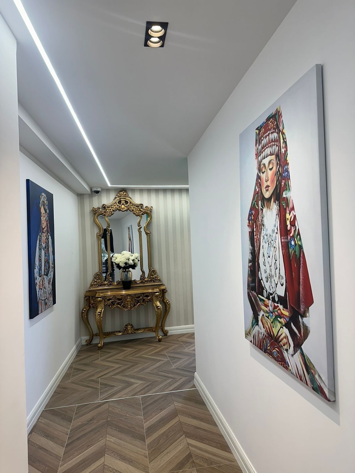 A hallway with a herringbone floor, a decorative mirror, and two paintings on the walls.