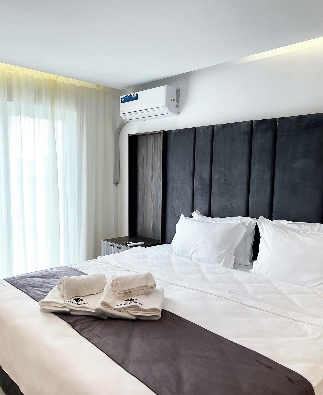 Une chambre d’hôtel avec un grand lit, du linge de lit blanc, une tête de lit sombre, un climatiseur, une table de chevet et une fenêtre avec des rideaux légers.
