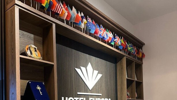 Een receptiebalie bij HOTEL EUROPA met een modern design, bestaande uit een houten balie, een grote presentatie van nationale vlaggen en een opvallend hotel logo.