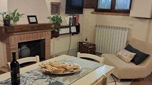 A cozy living room with a fireplace, a dining table with chairs, a sofa, and a television mounted on the wall. - The Sixth Moon Guest House in Pienza (Pienza)