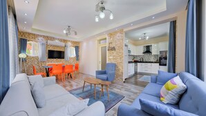 A modern living room with a sofa, armchairs, and a dining area with orange chairs. A kitchen is visible in the background. - Villa Maya (Fethiye)