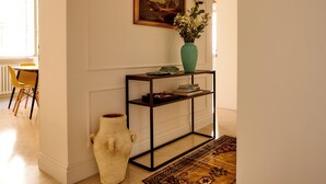 Ein Korridor mit weißer Wand, einem Gemälde, einer Blumenvase, einem Beistelltisch mit Büchern und einem gemusterten Teppich.