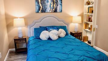 A bedroom with a blue bedspread, two bedside lamps, a white headboard, and a wall-mounted painting.
